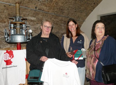 Dank an die Gastgeber: Übergabe vom Slow-Food-Genussführer im Gewölbekeller Drei Frauen in einem Gewölbekeller neben einer alten Spindelpresse, eine hält ein Slow-Food-T-Shirt, eine andere den Slow Food Genussführer.