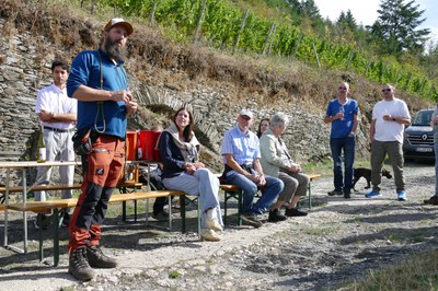 Verkostung am Fuß des Weinbergs. Gäste mit Weingläsern auf einem Schotterweg unterhalb eines Steillagen-Weinbergs mit Einzelpfählen.