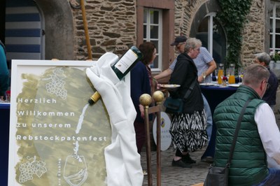 Willkommen zum Rebstockpatenfest Bemaltes Begrüßungsschild „Herzlich Willkommen zu unserem Rebstockpatenfest“ mit Weinflasche, dahinter Gäste im Hof.