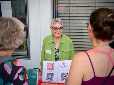 Eine Frau in grüner Jacke steht hinter einem Infotisch mit QR-Codes von Slow Food München und dem Genussführer 2027/28, davor zwei Besucherinnen