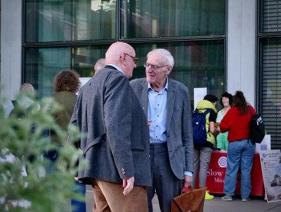 Zwei ältere Männer im Sakko im Gespräch, dahinter der rote Stand von Slow Food München und ein Aufsteller des Slow Mobils.