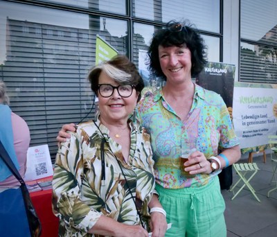 Zwei lächelnde Frauen, eine mit Bierglas, vor dem Slow Food München und Kulturland Genossenschaft Stand