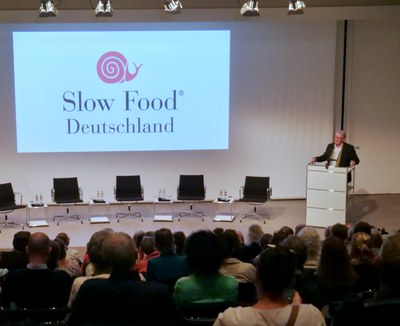 Ein Mann spricht am Rednerpult, daneben das Logo von Slow Food Deutschland auf der Leinwand, im Vordergrund das Publikum.