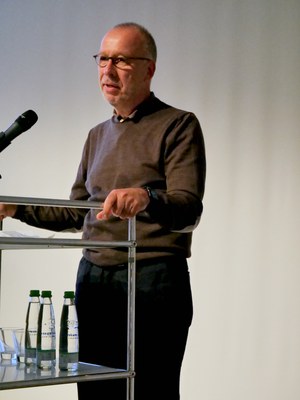 Ein Mann mit Brille und braunem Pullover spricht an einem Rednerpult ins Mikrofon.