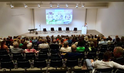 Hörsaal von oben mit vollen Sitzreihen, vorne eine Person am Rednerpult, daneben fünf leere Sessel und eine Projektion.
