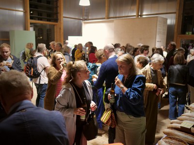 Viele Gäste stehen in einer Halle aus Holz im Gespräch, einige mit Bierflaschen, im Vordergrund Brotlaibe.