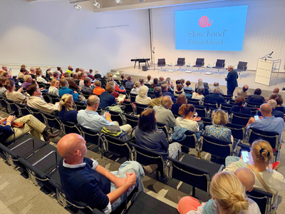 Voll besetzter Hörsaal mit ansteigenden Sitzreihen, auf der Leinwand das Logo von Slow Food Deutschland, auf der Bühne fünf leere Sessel und ein Keyboard.