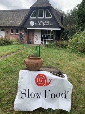 Forellenhof Benecke war Gastgeber des diesjährigen Tages der Fische 2026 © Slow Food Deutschland Reetgedecktes Gebäude der Forellenzucht Benecke mit Slow-Food-Banner im Vordergrund