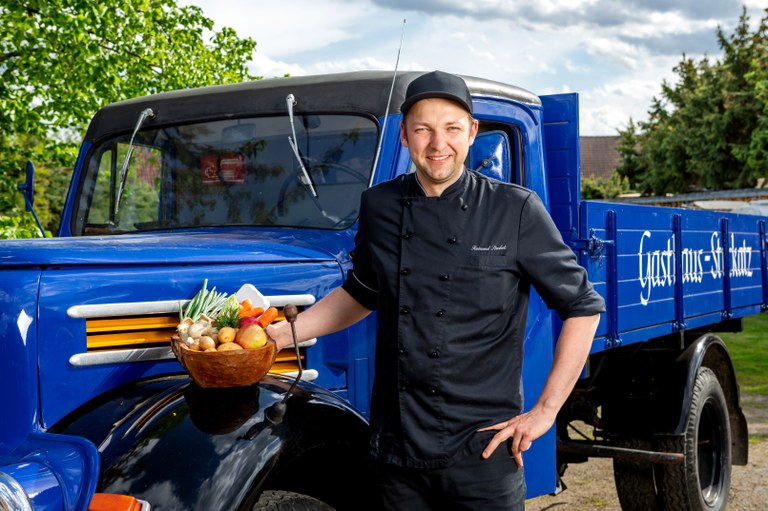 Raimund Stuckatz steht lächelnd vor einem blauen Oldtimer-Lieferwagen mit der Aufschrift Gasthaus-Stuckatz, in der Hand ein Korb mit regionalem Gemüse