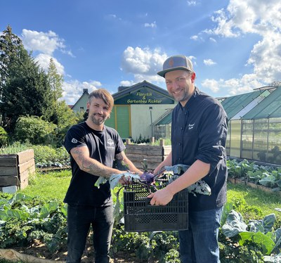 Raimund Stuckatz und ein weiterer Mann halten gemeinsam eine Kiste mit frisch geerntetem Kohlrabi in der Gärtnerei Richter