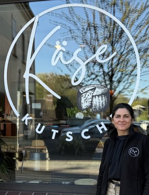 Yasemin Çelik vor ihrem Spezialitätenladen 'Käsekutsche' in Willich-Anrath (c) Yasemin Çelik Yasemin Çelik steht lächelnd in schwarzer Jacke vor dem Schaufenster der Käsekutsche in Willich-Anrath, auf dem Fenster das Logo mit Käselaib