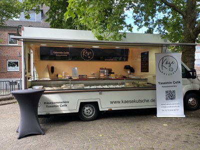 Der Marktwagen der Käsekutsche (c) Yasemin Çelik Weißer Verkaufswagen der Käsekutsche mit geöffneter Theke voller Käse auf einem Wochenmarkt, daneben ein Rollup mit dem Logo