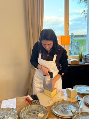 Käse schneiden, probieren, erklären (c) Yasemin Çelik Yasemin Çelik in weißer Schürze schneidet mit einem großen Käsemesser einen Käselaib an, davor Teller mit Käseproben