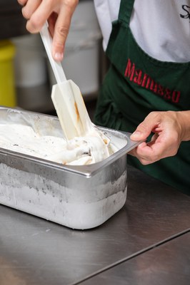 Hände streichen frisch gerührtes Eis aus einer Metallwanne, im Hintergrund Manusso-Schürze